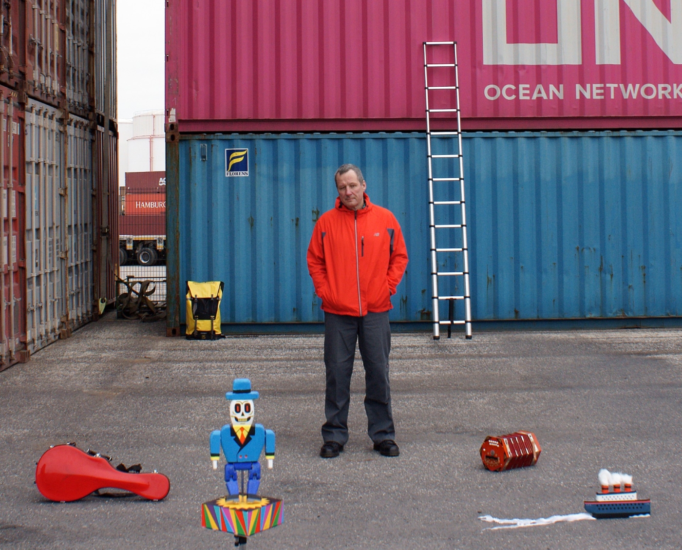 Der Bühnenbilder steht vor Frachtcontainern mit Objekten vor sich am Boden.